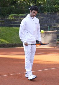 Balta sportinė striukė su užtrauktuku ir logotipu, su derančiomis kelnėmis. Vyras laiko teniso raketę, stovėdamas ant molio aikštelės.