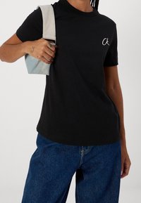 Černé bavlněné tričko s kulatým výstřihem a krátkými rukávy. Na prsou je malé vyšívané logo v bílé barvě. Kombinováno s modrými denimovými kalhotami.