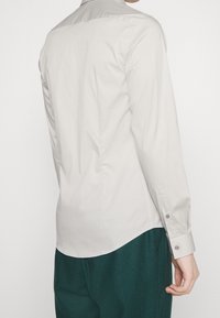 Camisa de botones gris claro de manga larga, con textura suave y dobladillo curvado. Presenta un cuello clásico y puños abotonados.