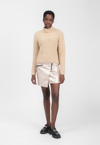 Kvindeklædt i en beige fuzzy sweater, en metallisk beige asymmetrisk miniskirt med lynlåsdetaljer og sorte loafers med guld-kæde accenter står imod en hvid baggrund.