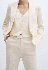 Costume en lin beige clair comprenant un blazer ajusté avec des revers crantés et un gilet boutonné. Le pantalon a une coupe cintrée et des poches latérales.