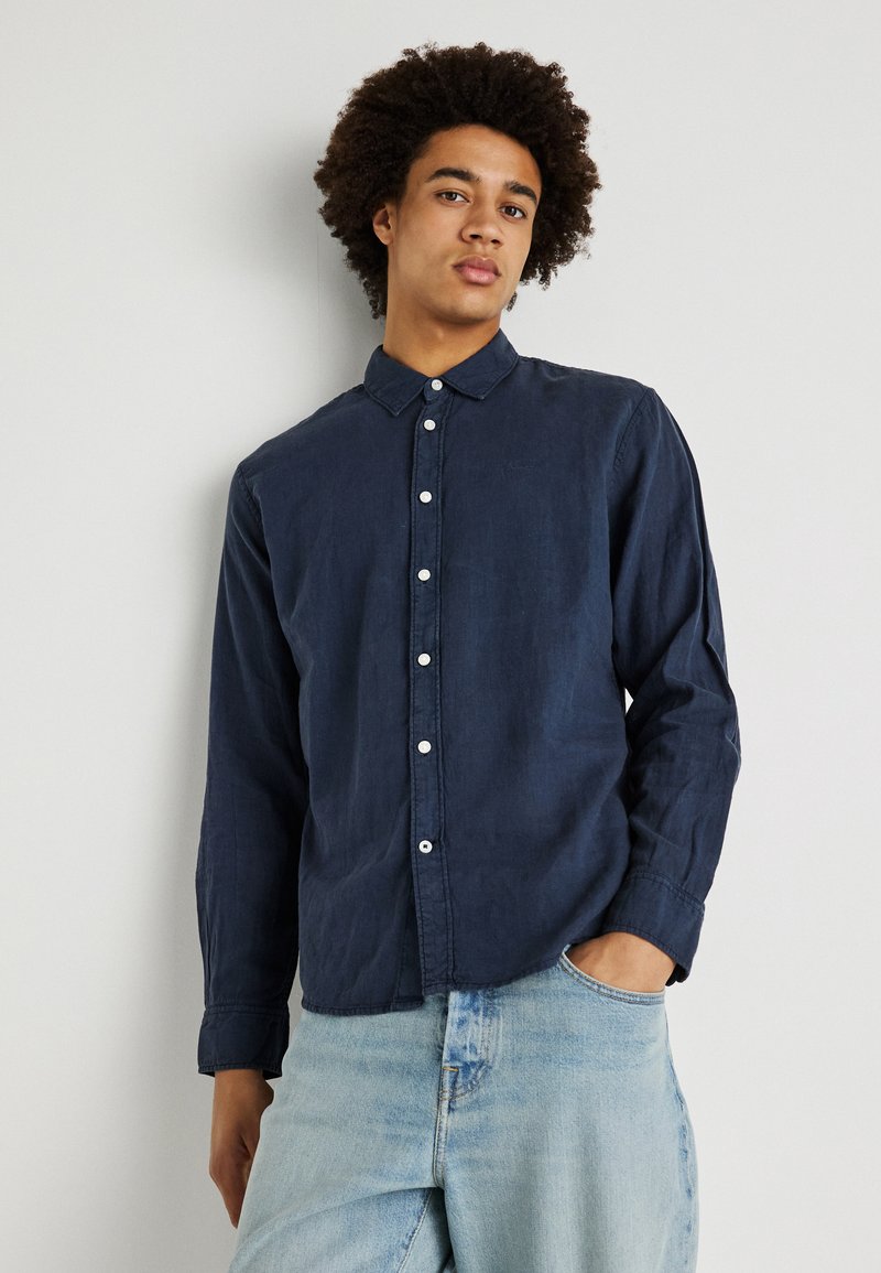 Camisa de botones azul marino hecha de tela texturizada, con cuello extendido y mangas largas; combinada con jeans de mezclilla azul claro.