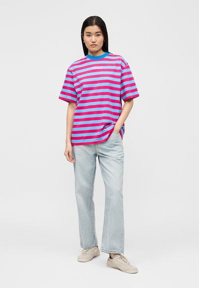 Γυναίκα που στέκεται με το χέρι στην τσέπη, φορά μπλε και ροζ ριγέ μπλουζάκι, ανοιχτόχρωμο τζιν παντελόνι και μπεζ αθλητικά παπούτσια σε μονόχρωμο φόντο.