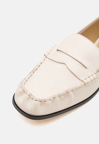 Beige leren loafers met een soepele textuur, voorzien van een gestikte vamp en een decoratief uitgesneden accent. Zwarte buitenzool zorgt voor contrast.