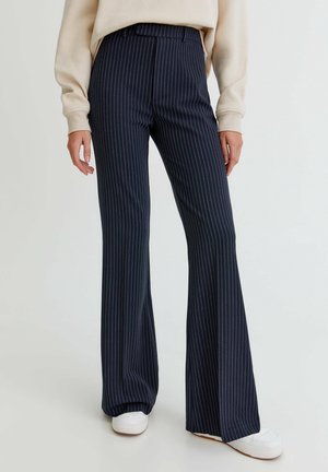 Woman wearing navy blue pinstripe wide-leg trousers paired with white sneakers and a beige long-sleeve top, standing on white floor.