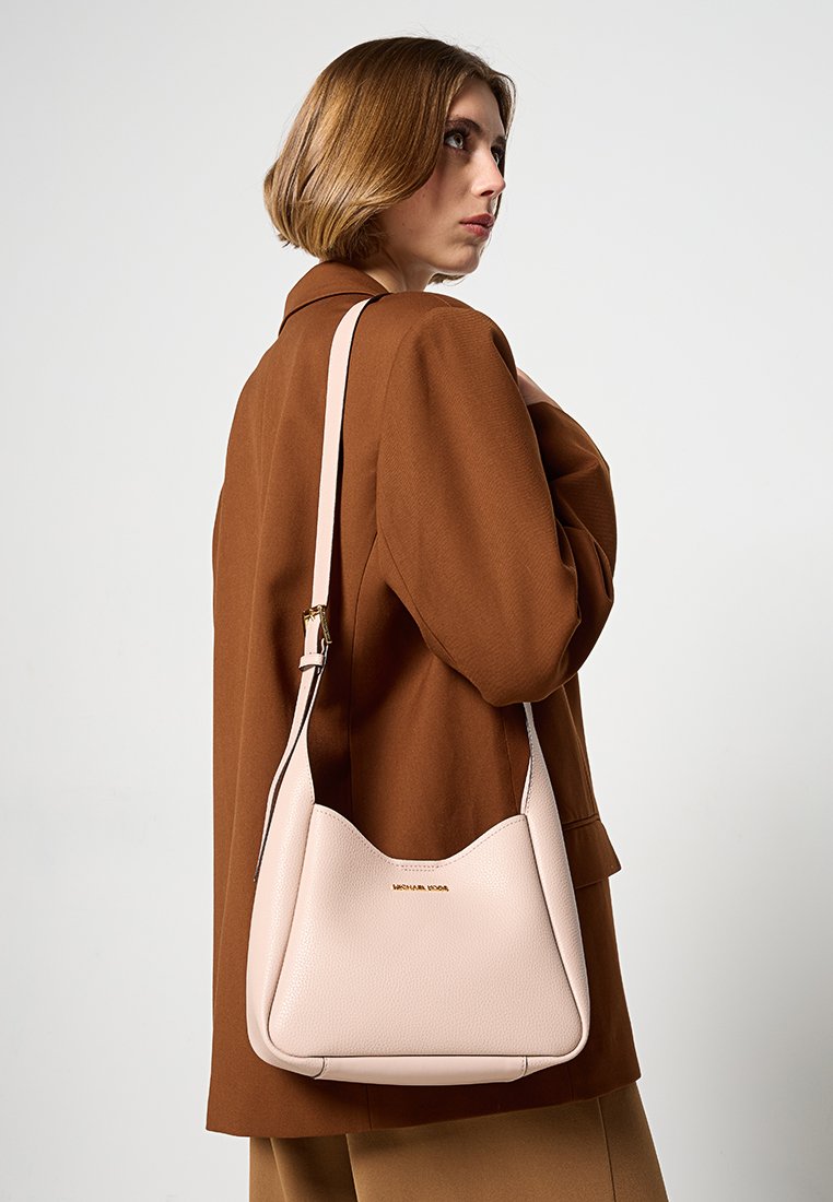 Sac à bandoulière en cuir rose pâle avec une forme arrondie et un logo doré, associé à un blazer oversize marron sur un fond neutre.