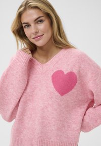 Pull rose en maille douce et texturée avec un motif en forme de cœur rose foncé contrastant sur la poitrine gauche et des poignets côtelés.