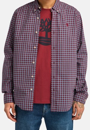 Mand iført en åben rød- og blåternet skjorte over en mørkerød t-shirt med sort trælogo, kombineret med blå jeans.