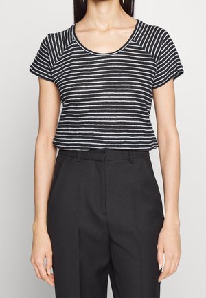 Kvinde iført en sort-hvid stribet kortærmet top, stoppet ned i højtaljede sorte bukser, foran en ensfarvet baggrund.