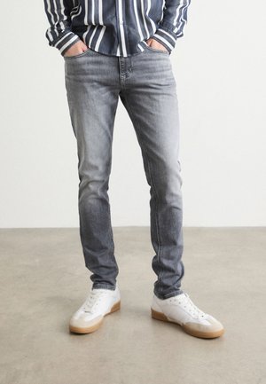 Mężczyzna w wyblakłych szarych obcisłych jeansach, białych sportowych butach z gumową podeszwą oraz w czarno-białej, paskowanej koszulce z długim rękawem i rękami w kieszeniach.