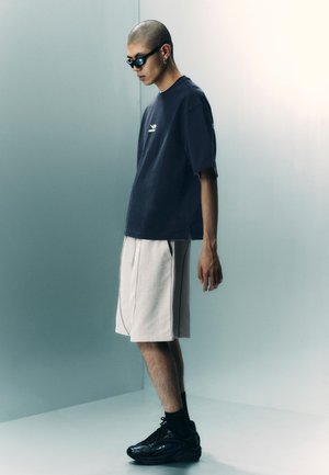Młody mężczyzna z ogoloną głową, noszący ciemne okulary przeciwsłoneczne, granatową, luźną koszulkę, białe szorty, czarne skarpety i czarne trampki, stojący w minimalistycznym otoczeniu.