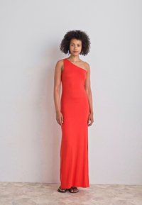 Raudona, vieno peties maxi suknelė, pagaminta iš lygaus, tamprus audinio su šonine iškarpa. Modelis stovi ant plytelėmis padengto grindinio prieš paprastą foną.