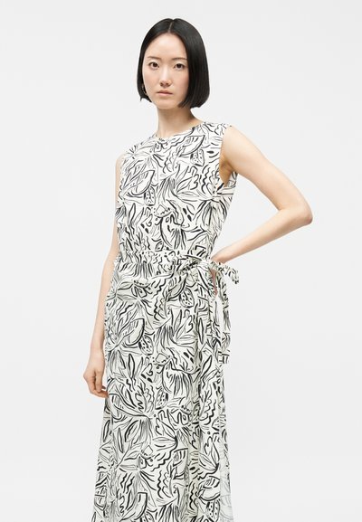Γυναίκα με κοντά μαύρα μαλλιά, φορεί λευκό αμάνικο φόρεμα με μαύρο αφηρημένο σχέδιο φύλλου και ζώνη που δένει στο πλάι, στέκεται μπροστά σε απλό φόντο.