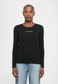 Vrouw in een zwart shirt met lange mouwen met het witte "EA7 Emporio Armani" logo en lichtblauwe jeans, staand tegen een effen witte achtergrond.