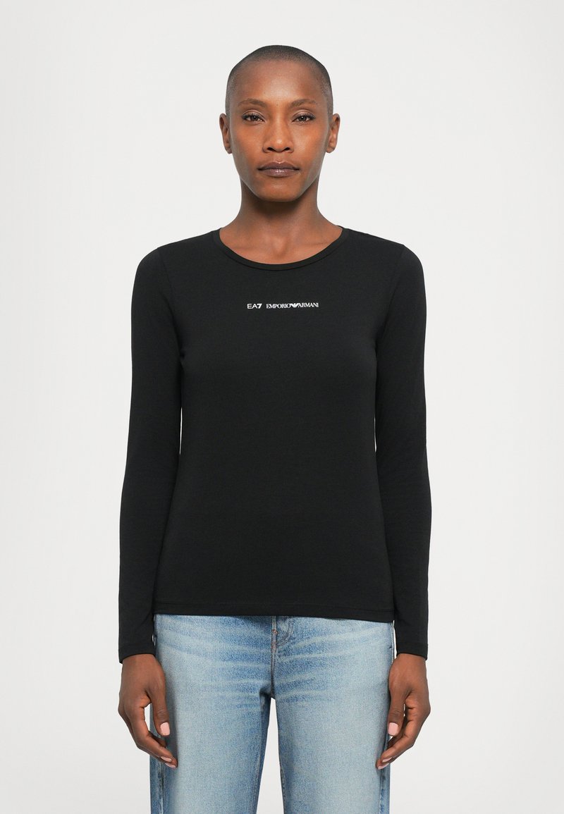 Vrouw in een zwart shirt met lange mouwen met het witte "EA7 Emporio Armani" logo en lichtblauwe jeans, staand tegen een effen witte achtergrond.