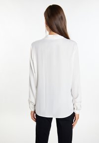 Bluză albă cu mâneci lungi, cu o textură netedă, guler înalt și manșete cu nasturi, asortată cu pantaloni negri. Design simplu și elegant.