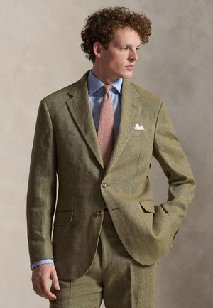 Άνδρας με σγουρά μαλλιά, ντυμένος με ανοιχτό καφέ κοστούμι, μπλε πουκάμισο, ροζ γραβατα με σχέδια και άσπρο μαντηλάκι στην τσέπη, στέκεται μπροστά σε μια απλή φόντο.