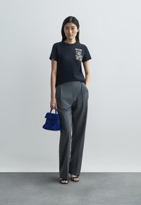 Μαύρο μπλουζάκι με γραφικό Moschino, γκρι ραμμένα παντελόνια και μια μικρή μπλε τσάντα. Το μοντέλο φορά μαύρες ψηλές γόβες.