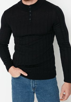 Uomo che indossa un maglione nero a coste con quattro bottoni sul colletto e jeans blu, in piedi con una mano in tasca.