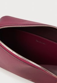 Borsa in pelle bordeaux con chiusura a zip e hardware in argento. L'interno presenta una texture liscia con il marchio "Calvin Klein" in rilievo.