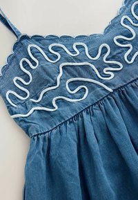 Denimklänning med blå nyans, intrikat vitt broderat mönster och plisserad kjoldesign med tunna remmar och vågiga kanter.