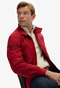 Rode zip-up jack met een hoge kraag, voorzien van een groen logo-opdruk op de linkermouw en geribbelde manchetten. Draagt over een beige trui.