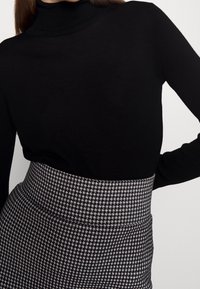 Femme portant un pull col roulé noir rentrée dans une jupe crayon à carreaux en noir et blanc à taille haute, la main posée sur la hanche.