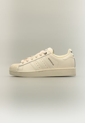 Klassischer cremefarbener Adidas Superstar Sneaker mit Zehenkappe, drei Streifen und Text "SUPERSTAR" an der Seite, im Seitenprofil gezeigt.
