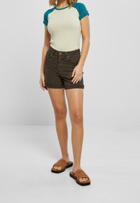 Lyhythihainen, kermavärinen t-paita, jossa on teal-värisiä yksityiskohtia, yhdistetty tiukkoihin tummanruskeisiin shortseihin. Asusteena on teksturoitu ruskea slaidi-sandaalit.