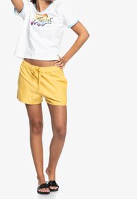 Femme portant un crop top blanc à manches courtes avec un texte coloré, un short jaune à cordon de serrage et des sandales noires tressées, debout avec la main sur la hanche.