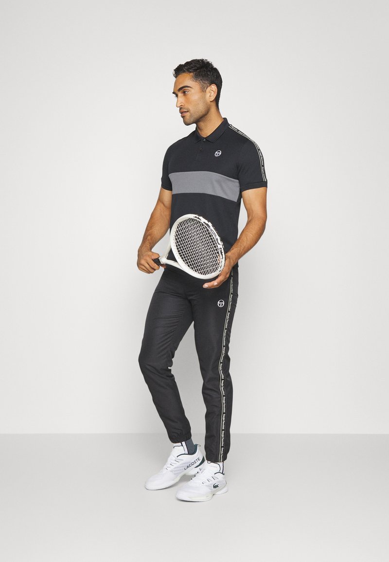 Polo noir avec rayure grise, pantalon de sport noir avec accents sur les côtés, tenant une raquette de tennis, portant des chaussures de sport blanches.