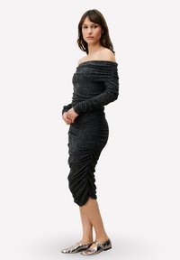 Robe noire mi-longue épaule dénudée avec texture froncée ; manches longues ; silhouette ajustée ; associée à des chaussures plates métalliques.