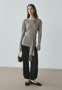 Szary długi rękaw z wiązaniem w talii, zestawiony z czarnymi spodniami o szerokich nogawkach i czarnymi płaskimi butami. Modelka trzyma małą brązową torebkę.