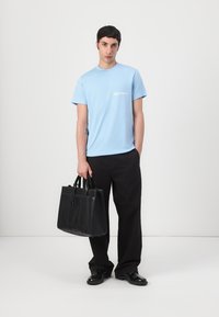 T-shirt en coton bleu clair à manches courtes, arborant un petit logo blanc, associé à un pantalon large noir et un sac fourre-tout en cuir noir.