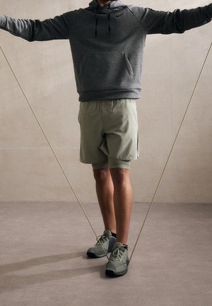 Person trägt grauen Kapuzenpullover und hellgrüne Shorts und benutzt Widerstandsbänder, während sie auf einem strukturierten Boden vor einer schlichten Wand steht.