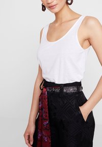 Haut blanc sans manches en tissu doux, associé à un pantalon noir à motifs avec une ceinture mise en valeur par un foulard coloré.