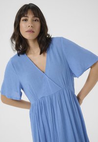 Helles blaues Kleid mit kurzen Ärmeln, V-Ausschnitt, strukturiertem Stoff, plissiertem Rock und dezentem, erhobenem Punkte-Muster.