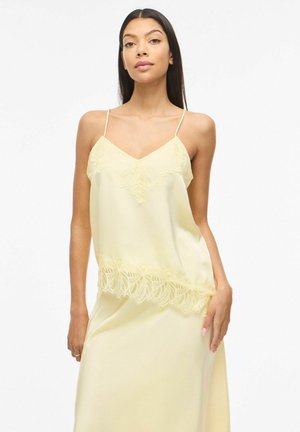 Kobieta ubrana w bladożółty satynowy kamizelowy top z koronkową lamówką i pasującą spódnicę, stojąca na jednolitym białym tle.
