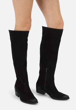 Schwarze kniehohe Stiefel mit niedrigen Absätzen und seitlichen Reißverschlüssen, getragen an nackten Beinen vor einem schlichten hellen Hintergrund.