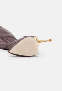 Burgundy-kuvioinen nahkakenkä, jossa on korkea korko ja terävä kärki, varustettuna kultasävyisellä stiletto-korkolla ja kumipintaisella, uurretulla pohjalla pidon parantamiseksi.