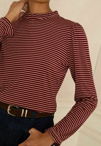 Bordozsarkana un krēmkrāsas svītrains garām piedurknēm krekls ar volānu apģērba kakla izgriezumā un aprocēm, valkāts kopā ar brūnu jostu un tumši dzeltenām džinsa biksēm.