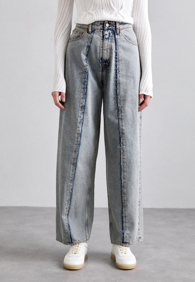 Jeans bleu clair à jambes larges avec une taille haute, présentant des accents usés et des coutures bien visibles. Associés à des baskets blanches.