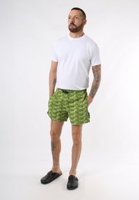 Pantaloni corti verdi con un motivo testuale nero ripetitivo, vita elastica e tasche laterali. Abbinati a una maglietta bianca semplice e zoccoli neri.