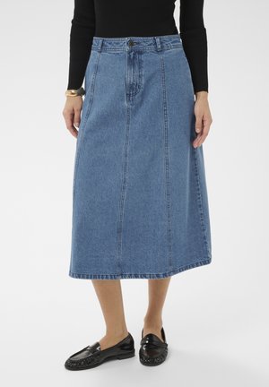 Kobieta ubrana w niebieską dżinsową spódnicę midi, czarną bluzkę z długim rękawem, czarne skórzane loafersy i złotą bransoletkę, stojąca na białym tle.