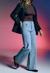 Femme portant un blazer noir, un haut marron ajusté, un jean bleu clair à jambes larges et des chaussures noires, marchant devant un fond dégradé rose et bleu.