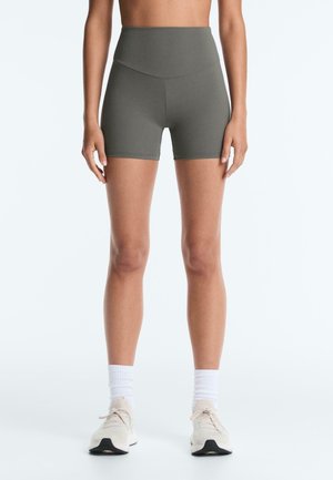 Kvinde iført olivengrønne atletiske shorts med høj talje, hvide crew-sokker og beige løbesko, stående mod en ensartet hvid baggrund.