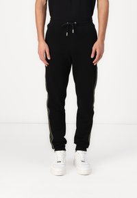 Schwarze Jogginghosen mit einem Kordelzugbund, versehen mit goldenen seitlichen Streifen und gerippten Bündchen. Kombiniert mit weißen Turnschuhen.