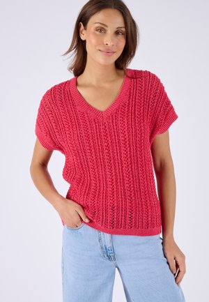 Donna che indossa un top lavorato a maglia rosa acceso con scollo a V e maniche corte, e jeans denim azzurri, in piedi su uno sfondo chiaro.