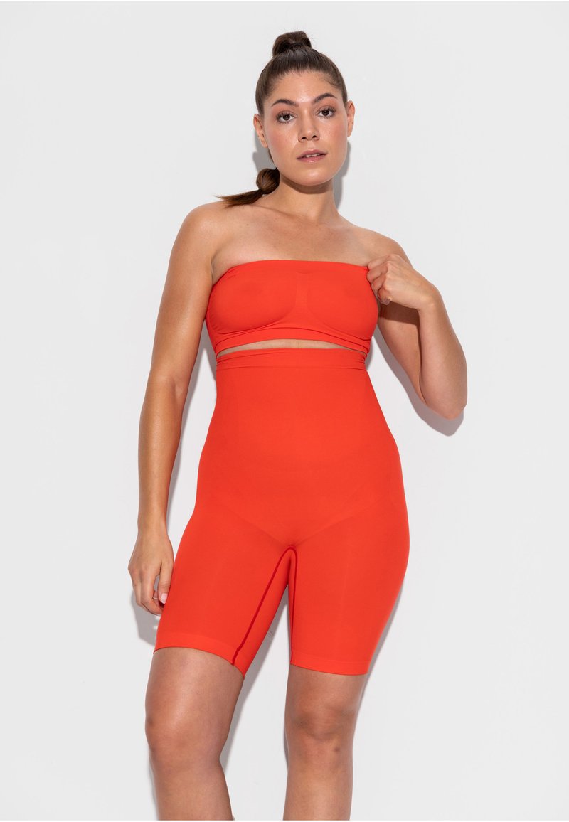 Moteris su tamsiais plaukais, surištais į aukštą uodegą, vilki raudoną, be petnešėlių trumpą viršutinį drabužį ir derančius aukšto liemens dviratininkų šortus, stovinti prieš baltą foną.