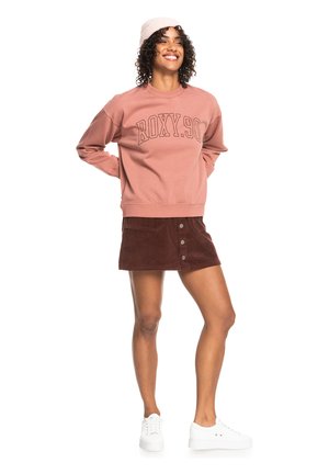 Ružový sweatshirt s nápisom "ROXY.90", hnedá kordová sukňa s prednými gombíkmi, biele tenisky a svetloružový pletený čiapka.